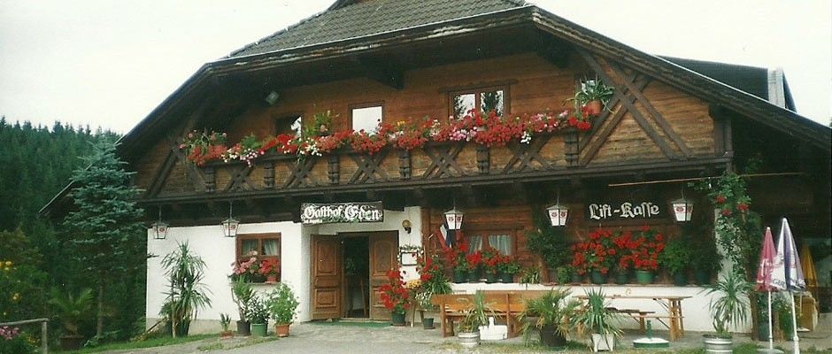 Das Äußere eines Holzchalets, 'Gipfel Stube', mit blumengeschmückten Balkonen, einer Holztür und einer Bank mit Topfpflanzen und Schirmen.