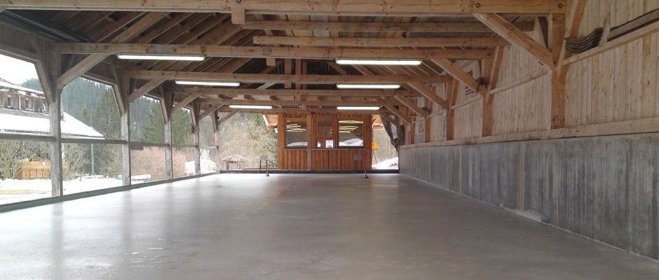 Eine leere, hölzerne Indoor-Eisbahn mit Holzbalken und Wänden, beleuchtet von Deckenleuchten, mit Blick auf das Freie durch große Glasfenster.