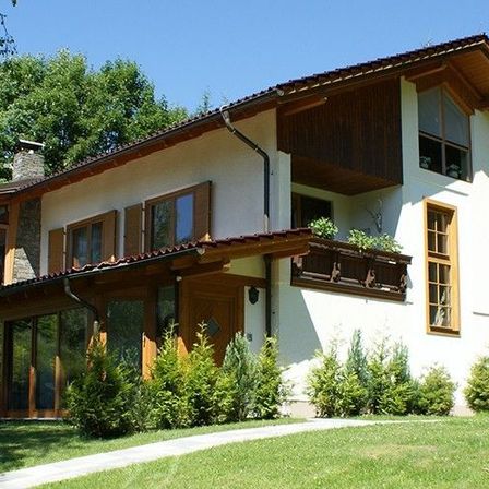 Ein großes Haus mit rotem Dach, umgeben von grünem Gras und Bäumen, mit einer Holzveranda, Glasfenstern und einem Balkon mit Pflanzen.