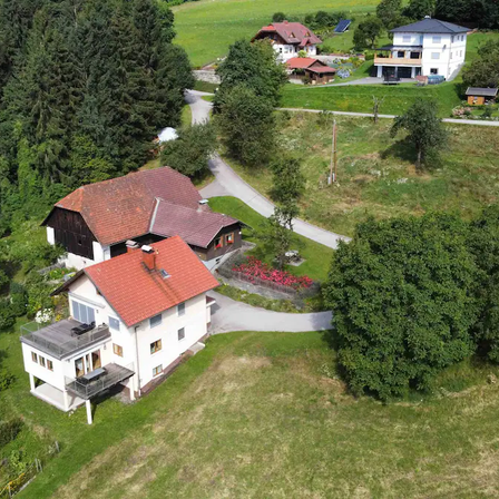 Luftaufnahme einer ländlichen Landschaft mit mehreren Häusern und grünen Wiesen. Die Häuser sind von Bäumen und Gärten umgeben.