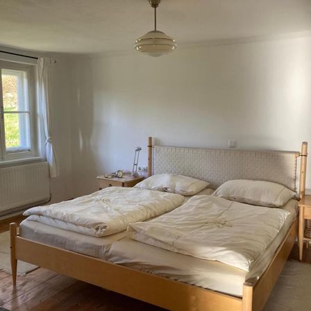 Ein Schlafzimmer mit einem Doppelbett und zwei Nachttischen auf beiden Seiten. Das Bett hat zwei Kissen, eine Lampe auf dem Nachttisch und einen Heizkörper unter dem Fenster.