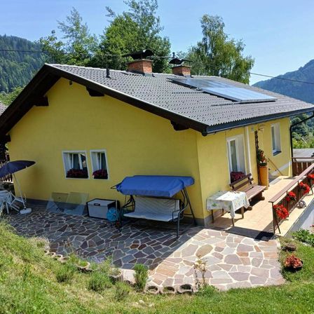 Ein gelbes Haus mit Solarmodulen auf dem Dach steht auf einem Hügel, umgeben von Grün und Bergen. Das Haus hat eine Bank, einen Tisch und einen Vordach auf der Veranda, mit einem Regenschirm und einer Aufbewahrungsbox in der Nähe.