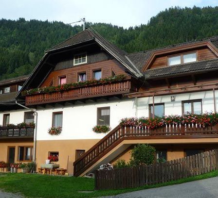 Ein zweistöckiges Haus mit einem braunen Dach, weißen Wänden und einem Holzzaun vorne. Das Haus hat einen Balkon mit roten Blumen im zweiten Stock.