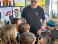 Ein Mann mit Brille hält eine Kamera, während eine Gruppe junger Mädchen ihn ansieht. Sie sind in einem Klassenzimmer mit Gemälden an der Wand.