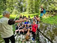 Eine Gruppe von Kindern und Erwachsenen sitzt auf Stufen im Wald, ein Mann zeigt auf einen Hund und ein Mädchen hält einen Becher.