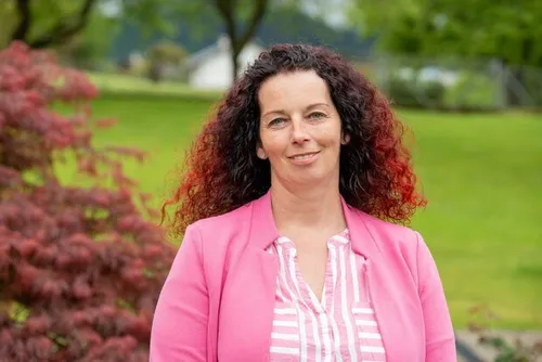 Eine Frau mit lockigem rotem Haar steht draußen und lächelt in die Kamera. Sie trägt eine rosa Jacke und ein weißes gestreiftes Hemd. Hinter ihr befindet sich ein grünes Grasfeld, einige Bäume und ein weißes Haus.