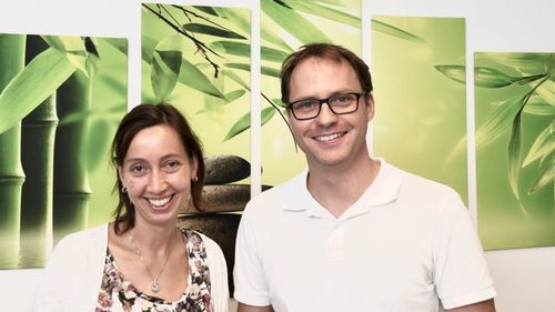 Ein lächelnder Mann und eine Frau stehen nebeneinander vor einer Wand mit grünen Blättern und Steinen.