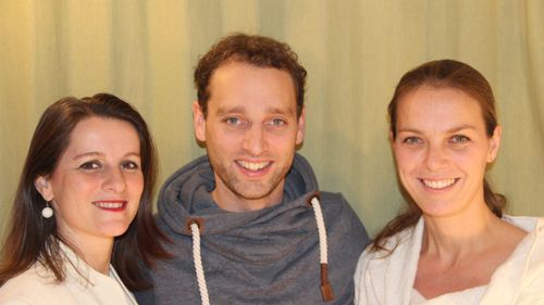Ein Mann in einer grauen Kapuzenjacke lächelt für ein Foto mit zwei Frauen, eine auf jeder Seite, alle vor einem hellgrünen Hintergrund.