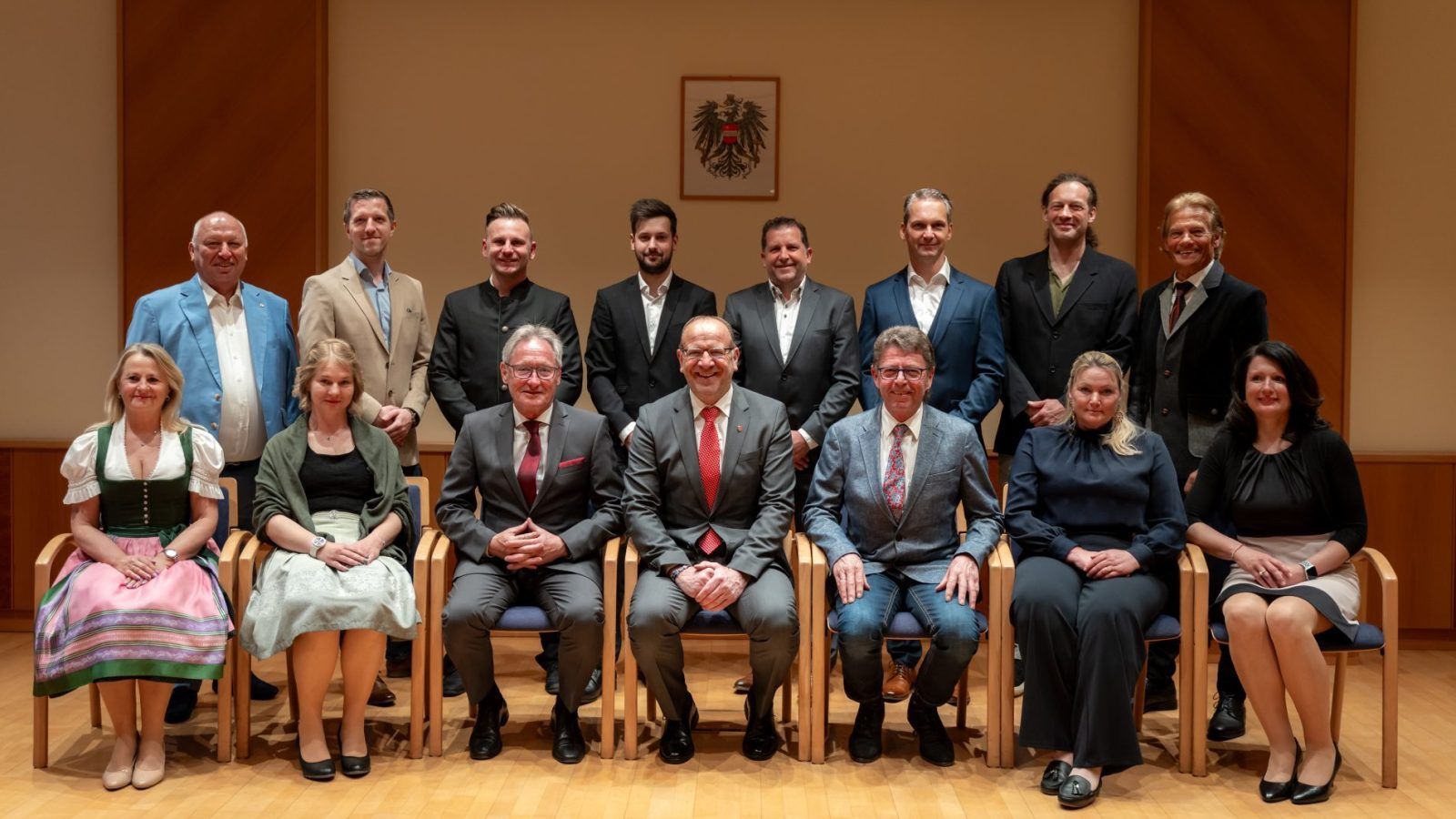 Eine Gruppe von Personen in formeller Kleidung, sowohl Männer als auch Frauen, sitzt und steht und posiert für ein Foto in einem Raum mit Holzböden und einer Wand mit einem gerahmten Bild.