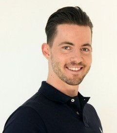 Ein Mann mit Bart und Schnurrbart lächelt fröhlich und trägt ein schwarzes Poloshirt mit einem einzelnen Knopf am Halsausschnitt. Sein Haar ist ordentlich gestylt.