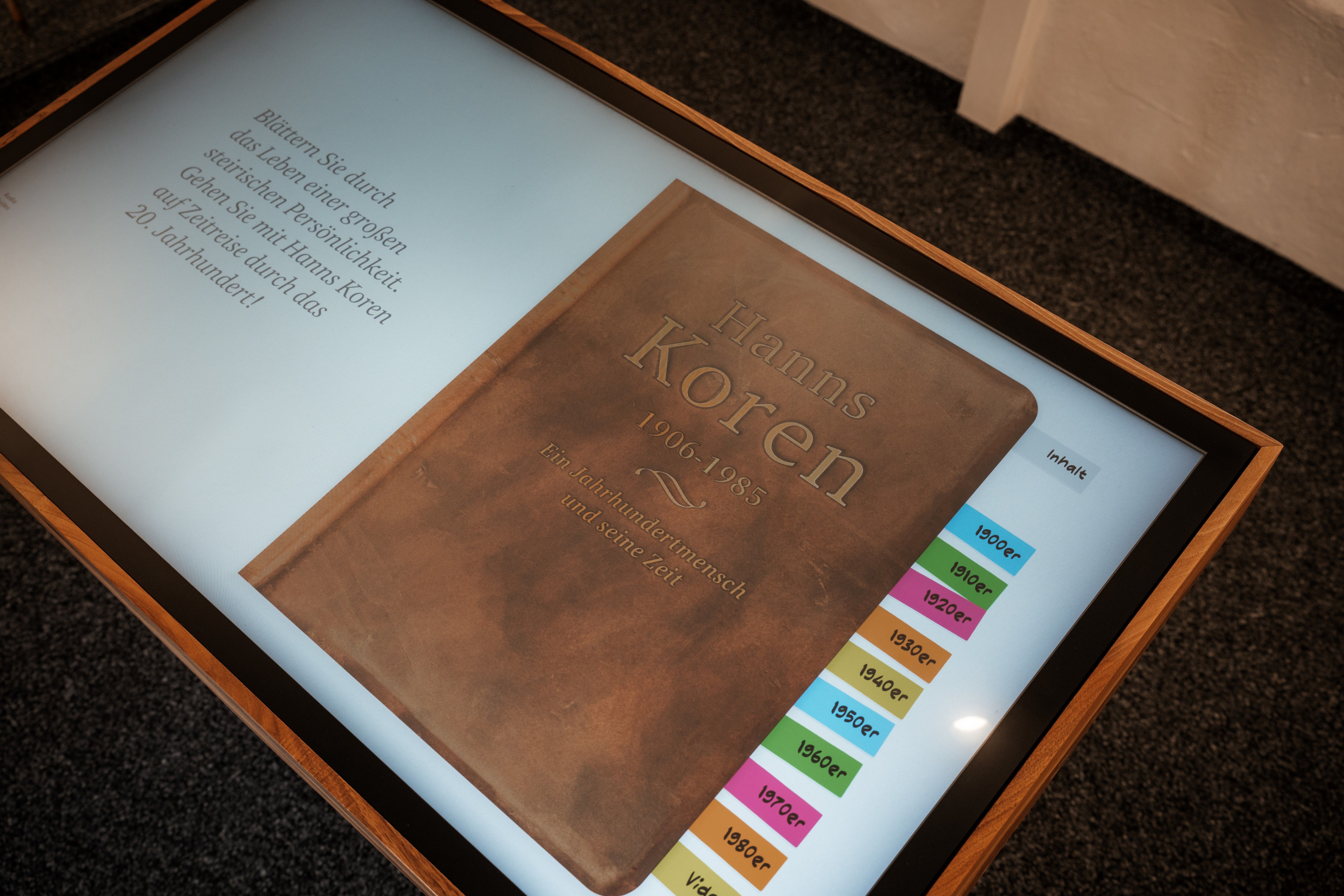 Ein braunes Buch mit dem Titel 'Hanns Koren' und den Jahren 1900 bis 1980 wird auf einem digitalen Bildschirm angezeigt. Das Buch ist in einem Holzrahmen montiert.