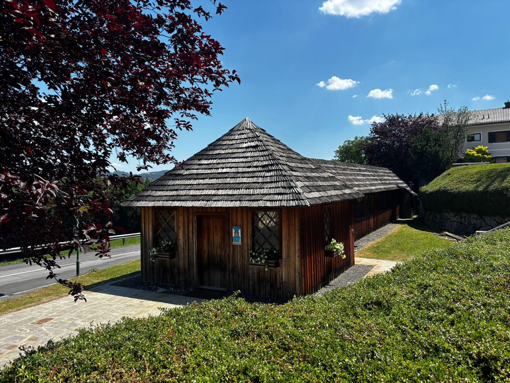 Ein kleines Holzhaus mit einem Strohdach und zwei Fenstern mit weißen Blumen. Es ist von Büschen und Gras umgeben, mit einer Straße in der Nähe.