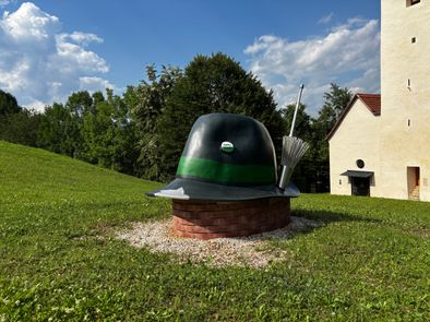 Ein großer Hut sitzt auf einem Ziegelsockel in einem Grasfeld mit einem Haus und Bäumen im Hintergrund.
