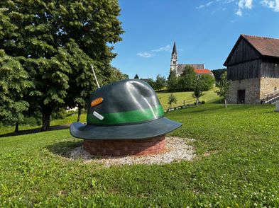Ein großer Hut steht auf einem Grashügel, mit einer Kirche und einem Gebäude im Hintergrund.