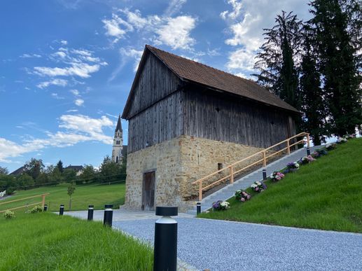 Ein Holzschuppen mit braunem Dach und Steinmauern. Stufen mit Handlauf führen zum Eingang. Es gibt Bäume, eine Kirche und Blumen um den Schuppen.
