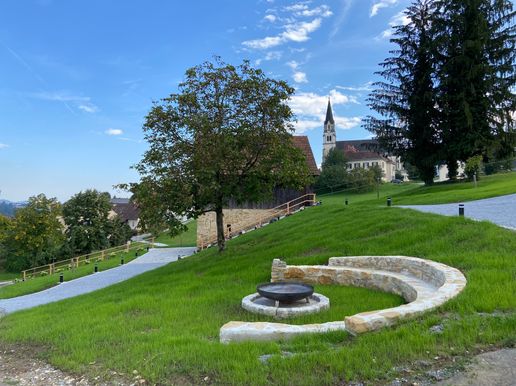 Ein grasbewachsener Hügel mit einem Baum und einer Steinsitzbank. Dahinter befindet sich ein Gebäude mit einem Turm. Der Himmel ist blau mit verstreuten Wolken.
