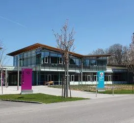 Ein modernes Gebäude mit Glaswänden und einem braunen Dach. Vor dem Gebäude befinden sich zwei pinkfarbene Schilder. Der Bereich vor dem Gebäude ist mit Gras und Bäumen bepflanzt.