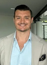 Ein lächelnder Mann mit hellblauem Hemd und hellgrauer Jacke mit schwarzem Taschentuch in der Tasche.