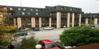 Ein mehrstöckiges Gebäude mit vielen Fenstern und einem Parkplatz davor. Mehrere Autos sind geparkt. Bäume und Sträucher befinden sich vor dem Gebäude.