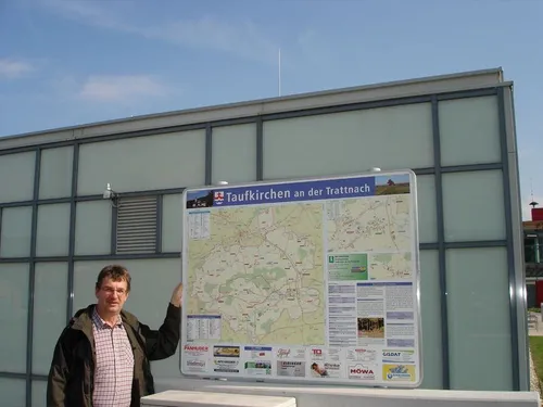 Ein Mann steht vor einem großen Kartenbrett und hält es fest. Das Brett trägt den Titel Taufkirchen an der Trattnach, mit einer Karte und weiteren Informationen.