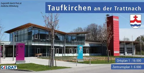 Ein Gebäude mit einem blauen Schild und einem roten Turm ist abgebildet. Es hat ein Logo darauf und eine lila Fahne. Bäume und ein Rasenbereich sind davor. Der Text ist auf Deutsch.