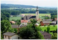 Eine Dorfkirche mit einem Turm ist von Häusern und Grünflächen umgeben. Die Landschaft ist üppig mit grünen Feldern und Bäumen.