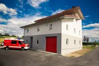 Ein roter Krankenwagen steht vor einem zweistöckigen Gebäude mit einer roten Garagentür. Der Himmel ist blau mit weißen Wolken.