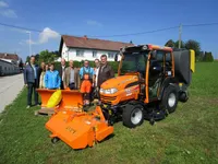 Eine Gruppe von Menschen posiert neben einem Traktor und einer Rasenmäher vor einem Haus.