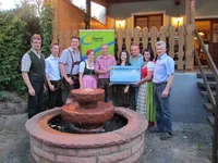 Eine Gruppe von Menschen steht vor einem Brunnen und hält einen großen Scheck. Sie sind in traditioneller deutscher Kleidung gekleidet. Die Szene spielt vor einem Holzhaus mit Treppen und Zaun.