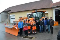 Eine Gruppe von Menschen steht vor einem orangefarbenen Traktor. Sie lächeln und scheinen für ein Foto zu posieren. Sie tragen Jacken und Hosen. Hinter dem Traktor steht eine Fahne. Der Traktor hat ein Namensschild mit der Aufschrift 'KAHLBACHER'. Ein Gebäude mit einer Garage befindet sich hinter ihnen.