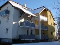 Ein gelbes und weißes Mehrfamilienhaus mit Balkonen und Schnee auf dem Dach. Das Gebäude hat drei Etagen mit Balkonen.