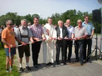 Ein Priester und mehrere Männer stehen auf einer Straße mit einem Bandschneider vor einer Menschenmenge.