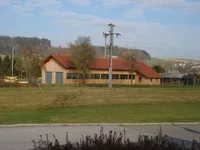 Eine große Scheune mit rotem Dach und Fenstern steht auf einem Grasfeld, mit einem Versorgungsmast in der Nähe. In der Ferne sind ein Haus und ein Wald zu sehen.