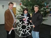 Drei Personen lächeln und posieren für ein Foto, während sie eine große Schneeflockendekoration halten. Hinter ihnen steht ein geschmückter Weihnachtsbaum.