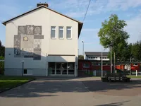 Ein großes weißes Gebäude mit einer Wandmalerei und Glastüren, einer Laterne und einer Bank. Das Gebäude hat drei Fenster und einen Schornstein. Bäume und ein Rasenbereich sind davor.