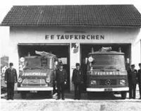 Ein Schwarz-Weiß-Foto zeigt Feuerwehrleute vor einer Feuerwache mit zwei Feuerwehrfahrzeugen. Die Wache ist mit 'FF Taufkirchen' beschriftet.