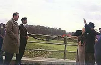 Eine Gruppe von Menschen steht in der Nähe eines Zauns, eine Person winkt. Dahinter befindet sich ein grasbewachsenes Feld.