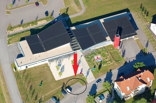 Eine Luftaufnahme eines Gebäudes mit einem roten Pfeil, der auf einen kreisförmigen Bereich, möglicherweise einen Parkplatz, zeigt. In der Nähe sind Autos geparkt, ein roter Pfeiler und ein Garten vor dem Gebäude.