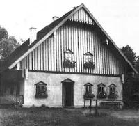 Ein schwarz-weißes Bild eines kleinen, traditionellen Hauses mit einem steilen Dach, Holzverkleidung und mehreren Fenstern mit Blumenkästen. Das Haus ist von Grün umgeben und hat eine Tür in der Mitte.