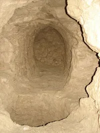 Ein schmaler, ovaler Tunnel, in die Erde gehauen, mit Wänden aus braunem, rauem Material. Der Tunnel führt nach unten, mit einem klaren Weg.