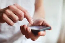 Eine Person hält ein Smartphone in beiden Händen. Sie scheint sich auf den Bildschirm zu konzentrieren, möglicherweise liest oder interagiert sie mit dem Gerät.