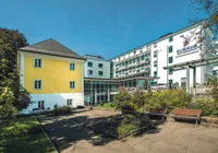 Ein großes weißes und gelbes Gebäude mit einem Garten davor, Klinikum steht an der Wand. Vor dem Garten stehen zwei Bänke.