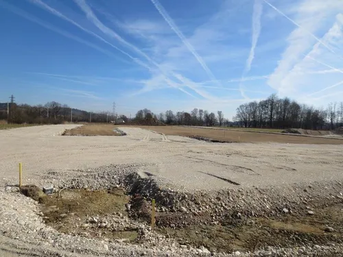 Ein leeres Baustellengelände mit einem Schotterweg, Haufen von Steinen und einem klaren blauen Himmel mit Wolkenspuren. Im Hintergrund sind Bäume und Stromleitungen zu sehen.