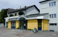 Ein Gebäude mit gelben Wänden und blauen Fenstern hat ein Schild über dem Eingang. Rechts sind eine Bank und ein Schild.