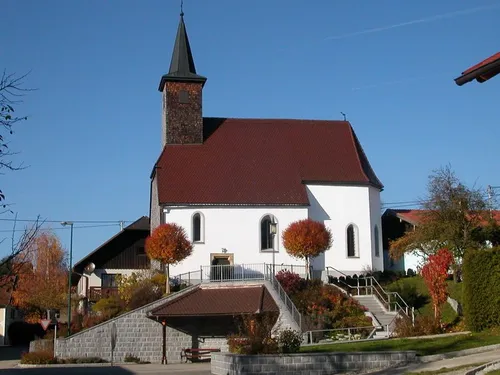 Eine weiße Kirche mit rotem Dach und Turm steht auf einem Hang. Es gibt Stufen, die zum Eingang führen, mit Bäumen und Pflanzen drumherum.