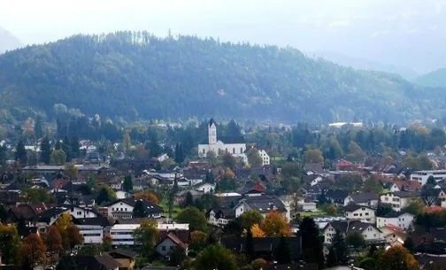 Eine Stadt mit vielen Häusern, umgeben von Bäumen und Bergen. Im Zentrum steht eine Kirche mit einem Turm.