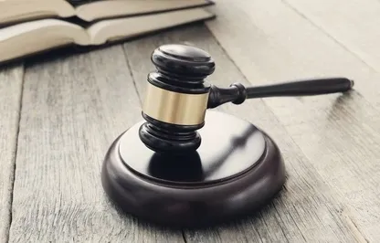 Auf einem Holztisch liegen ein hölzerner Gerichtshammer und ein offenes Buch. Der Hammer hat einen schwarzen Griff und einen goldenen Ring, während die Seiten des Buches weiß sind.