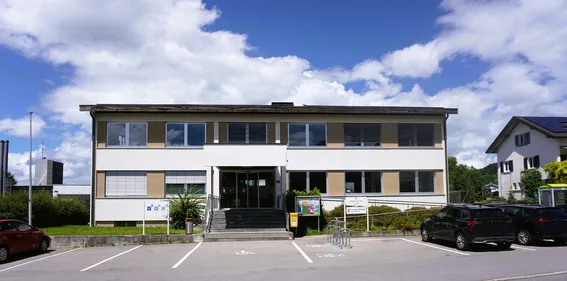 Ein Gebäude mit Glasfenstern und Türen, eine Treppe zum Eingang und einen Mülleimer auf der linken Seite. Ein Schild steht rechts und ein Auto ist vor dem Gebäude geparkt.