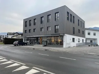 Ein modernes Gebäude mit Glasfenstern und einem geparkten Auto davor. Das Gebäude hat ein Schild oben und einen Bürgersteig mit einem Fahrradständer. Die Straße hat weiße Markierungen und ein dreieckiges Muster. Der Himmel ist bewölkt.