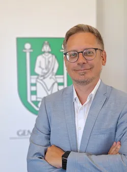 Ein Mann in einem hellblauen Anzug und mit Brille steht mit verschränkten Armen vor einem verschwommenen grünen Logo.